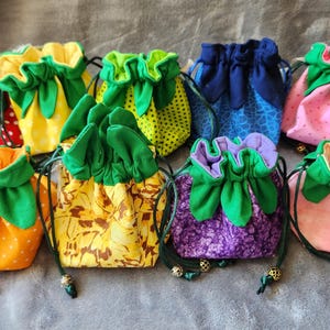 May include: A collection of drawstring pouches in various colors and patterns, resembling fruits. The pouches feature green leaf-like accents at the top and are made of fabrics with polka dots, floral designs, and other patterns. Each pouch has a drawstring closure.
