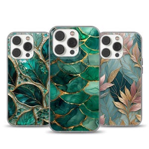 May include: Three phone cases with artistic designs. The cases feature leaf and abstract patterns in shades of green, gold, and pink. The designs are printed on clear cases, showcasing the phone's camera and buttons.