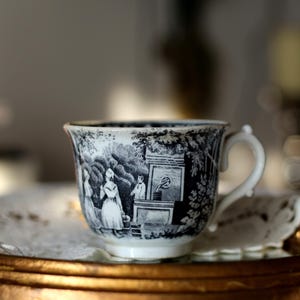 Könnte beinhalten: Eine weiße Keramiktasse mit einem schwarzen Transferdruck, der eine ländliche Szene mit Figuren an einem Steinbrunnen zeigt. Die Tasse verfügt über einen geschwungenen Henkel und einen ausgestellten Rand mit detaillierten Illustrationen.