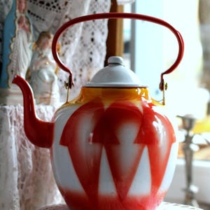 May include: A vintage enamel teapot with a red handle, spout, and decorative pattern. The teapot features a white body with a red and yellow design. The teapot is displayed on a lace doily.
