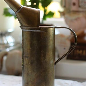 Könnte beinhalten: Vintage Ölkanne aus Messing mit Ausguss und Griff. Die Kanne hat einen zylindrischen Körper und einen gebogenen Griff. Der Ausguss ist nach oben abgewinkelt. Die Kanne ist gealtert, mit sichtbaren Gebrauchsspuren und Patina.