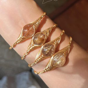 May include: Four wire-wrapped bracelets featuring round, translucent stones with internal golden needle-like inclusions. Each bracelet is crafted from gold-toned wire, twisted into a band with a decorative, pointed setting around the stone. Text reads ChicLliteCo.