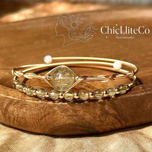 May include: A gold-toned wire bracelet featuring a central round rutilated quartz stone, a secondary strand of small translucent yellow beads, and two small white spherical end caps. The logo ChicLliteCo HandmadeJewelry is visible above.