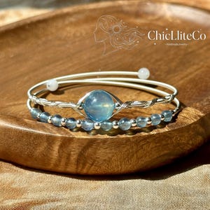 May include: A double-strand bracelet featuring a central light blue spherical bead wrapped in silver-toned wire, accompanied by a second strand of smaller translucent blue beads and silver spacers, with the text ChicLliteCo HandmadeJewelry.