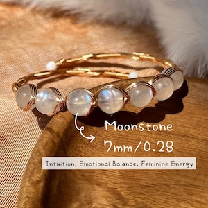 Op de afbeelding: Een goudkleurige draadarmband met zeven maansteen kralen, elk ongeveer 7 mm in diameter. De armband is voorzien van de tekst "Moonstone 7mm/0.28" en "Intuition, Emotional Balance, Feminine Energy."
