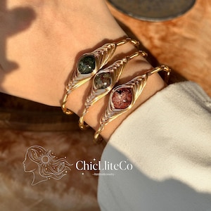 May include: Three gold-toned wire bracelets with gemstone accents are displayed on a wrist. Each bracelet features a different colored gemstone: green, brown, and red. The bracelets are handmade, as indicated by the text "ChicLiteCo - Handmade Jewelry".
