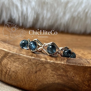 May include: A wire-wrapped ring featuring four round, dark blue, translucent beads secured within a gold-toned metal frame. The jewelry piece displays the text ChicLliteCo -HandmadeJewelry- above the beads, with a line art logo of a profile.