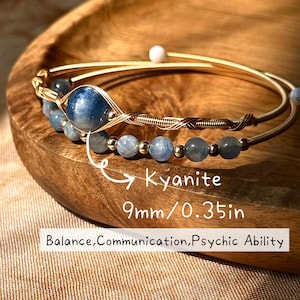May include: Two gold-coloured bracelets with blue kyanite beads and a larger blue kyanite stone. The bracelets are displayed on a wooden surface. The text on the image reads "Kyanite 9mm/0.35in Balance, Communication, Psychic Ability."