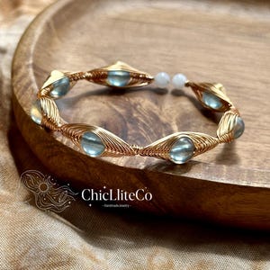 Op de afbeelding: Een goudkleurige, met draad omwikkelde armband met meerdere lichtblauwe, doorschijnende, ronde cabochonstenen. De armband heeft een open ontwerp met twee kleine witte kralen aan de uiteinden. De tekst ChicLiteCo -HandmadeJewelry- is zichtbaar.