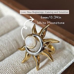 May include: A gold and silver ring with a white moonstone center, shaped like a sun and crescent moon. The ring is 0.24 inches in diameter and is displayed in a jewelry box. Text on the image reads: Love, New Beginnings, Calming and Intuition, White Moonstone.