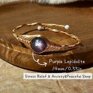 Könnte beinhalten: Ein drahtgewickeltes Armband mit einem zentralen 14 mm großen, violetten Lepidolith-Stein. Der goldfarbene Metalldraht bildet ein gedrehtes Band mit kleinen weißen Perlen an den Enden. Der Text lautet: Purple Lepidolite 14mm/0.55in Stress Relief & Anxiety&Peaceful Sleep.