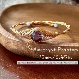 May include: A gold-colored wire-wrapped bracelet featuring a 12mm (0.47in) Amethyst Phantom stone. The bracelet has a woven design and two small white beads. Text on the image reads: Spiritual Transformation, Inner Growth, Wealth Manifestation.