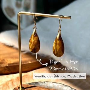 May include: A pair of teardrop-shaped Tiger's Eye earrings, each 0.9 inches long, displayed on a gold-colored stand. The earrings have a warm, golden-brown color with natural patterns. The text on the image says "Wealth, Confidence, Motivation."
