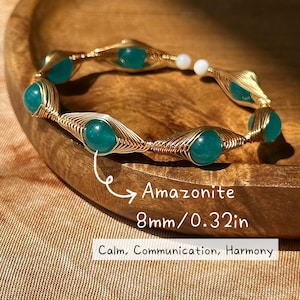 May include: A gold-toned wire bracelet featuring vibrant teal Amazonite beads, each 8mm (0.32in) in diameter, interspersed with small white beads. The bracelet is displayed on a wooden surface, with text indicating the Amazonite's properties: Calm, Communication, Harmony.