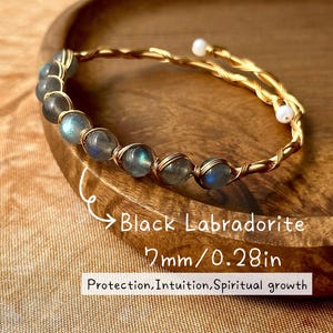 May include: A gold-toned wire bracelet with black labradorite beads. The bracelet features a twisted wire design and small white beads. The text on the image reads "Black Labradorite 7mm/0.28in Protection, Intuition, Spiritual growth."