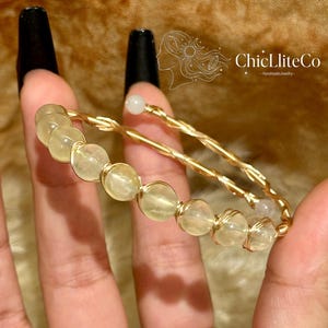 May include: A gold-toned wire-wrapped bracelet featuring a series of pale yellow, translucent spherical beads. The wire is twisted into a double-strand design, with small white beads at each end. The text ChicLiteCo HandmadeJewelry is visible.