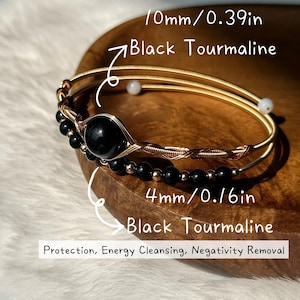 Pode incluir: Duas pulseiras de arame dourado com contas de turmalina negra. Uma pulseira apresenta uma pedra de turmalina negra de 10 mm (0,39 pol.) e a outra tem contas de 4 mm (0,16 pol.). O texto na imagem diz "Protection, Energy Cleansing, Negativity Removal."