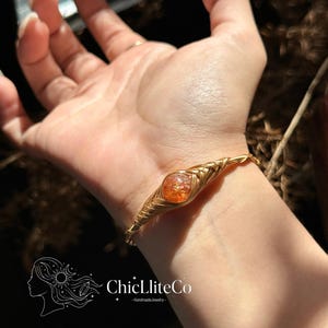 May include: A gold-toned wire-wrapped bracelet featuring a central round orange crackle glass bead. The metal wire is intricately coiled around the bead and extends into a thin band. The logo ChicLliteCo HandmadeJewelry is visible.