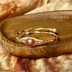 May include: A gold wire-wrapped bracelet featuring a central round orange sunstone and two small white beads at the ends. The metal wire is twisted into a spiral design. The text ChicLiteCo HandmadeJewelry is visible above the bracelet.