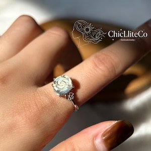 May include: A silver ring with a rose-shaped gemstone. The ring has a slender band and a small floral detail. The gemstone has an iridescent, pearlescent sheen. The brand name "ChicLliteCo" is visible in the upper right corner.