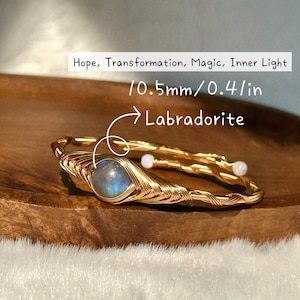 May include: A gold-colored wire-wrapped bracelet with a central labradorite stone and two small white beads. The labradorite is 0.4 inches in diameter. Text on the image reads "Hope, Transformation, Magic, Inner Light" and "Labradorite".