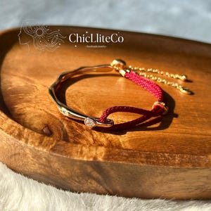 May include: A gold-toned bracelet with a red cord and a small, clear gemstone. The bracelet has a unique, wavy design and is displayed on a wooden tray. The brand name "ChicLiteCo" is visible in the background.