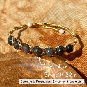 May include: A gold wire-wrapped bracelet featuring six dark grey beads, each 8mm or 0.32in in diameter, labeled as Iolite. The bracelet ends with two small white beads. Text on the image reads: Iolite, 8mm/0.32in, Courage & Protection, Intuition & Grounding.
