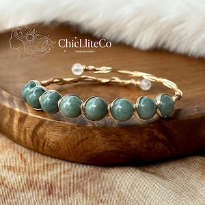 May include: A wire-wrapped bracelet featuring seven smooth, round, sage green beads secured with gold-toned wire. The bracelet has an open-ended design with two small, clear, spherical beads at the terminals. The text ChicLiteCo is visible.