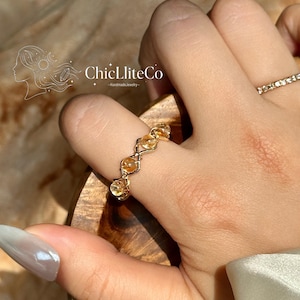 Può includere: Un anello in filo metallico dorato caratterizzato da una serie di piccole perle rotonde e traslucide color giallo citrino, disposte in un motivo geometrico ripetuto. L'anello è indossato su un dito, con una sottile fascia dorata su un altro.