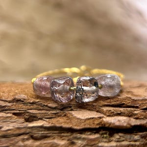 May include: A gold-toned ring featuring four clear, square-cut gemstones with internal inclusions. The gemstones are a mix of clear and pink hues. The ring is set on a wooden surface.