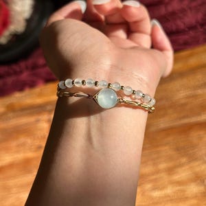 May include: Two bracelets are displayed. The top bracelet features small, white beads and gold-coloured accents. The bottom bracelet has a gold-coloured wire design with a large, round, light blue stone in the centre. The bracelets are on a person's wrist.