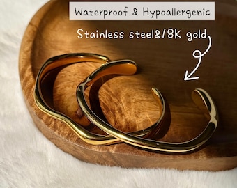 May include: Gold-coloured stainless steel and 8K gold bracelets. The open cuff bracelets are displayed on a wooden tray. The text on the image reads: Waterproof & Hypoallergenic.