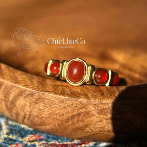 May include: A gold-toned ring with a central oval-shaped red gemstone, flanked by three smaller, round red gemstones. The ring is set on a wooden surface. The brand name "ChiclliteCo - handmade jewelry" is visible in the background.