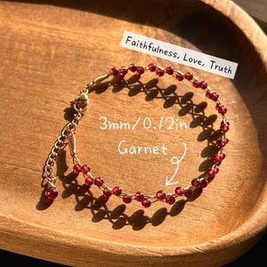 May include: A delicate garnet bracelet with 3mm beads, strung on gold wire. The bracelet is displayed on a wooden tray, with the words "Faithfulness, Love, Truth" written on a white label.
