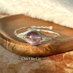 May include: A wire-wrapped bracelet featuring a round, translucent, light purple gemstone at the centre. The band is constructed from twisted silver-toned wire with small, round, white beads at the ends. The text ChicLliteCo is visible.