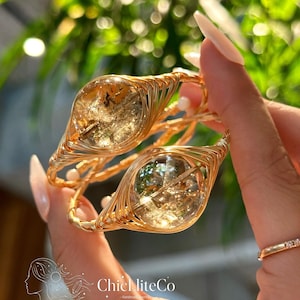 May include: Gold-toned wire-wrapped bracelets with clear, oval-shaped stones containing inclusions. The bracelets are held up against a blurred green background. The bracelets are handmade and feature the text "ChicLiteCo - handmade jewelry".