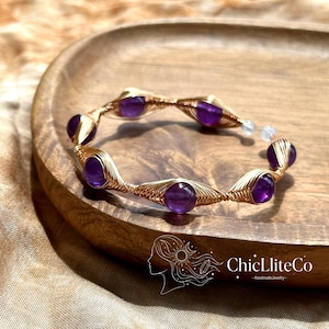 Può includere: Un bracciale a fascia in filo metallico avvolto con perline rotonde di ametista viola scuro, fissate all'interno di una struttura in filo metallico color oro. Il design include un motivo ripetitivo di segmenti di filo avvolti tra ogni perla. Il testo ChicLliteCo è visibile.