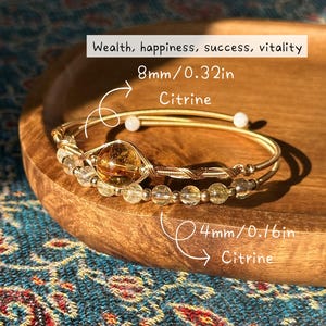 May include: A gold-toned bracelet with citrine beads and a larger citrine stone, 8mm/0.32in and 4mm/0.16in. The bracelet is displayed on a wooden tray with the text "Wealth, happiness, success, vitality".
