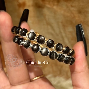 May include: Two wire-wrapped bracelets featuring round, dark gray, reflective beads. One bracelet is constructed with gold-toned wire, while the other uses silver-toned wire. The text ChicLliteCo -HandmadeJewelry- is visible in the center.
