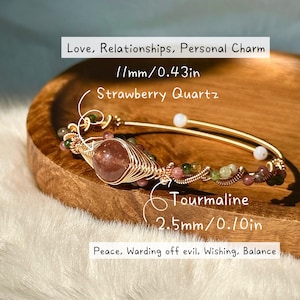 May include: A gold-colored wire-wrapped bracelet featuring a large strawberry quartz stone and smaller tourmaline beads. The bracelet is accented with additional small beads and is displayed on a wooden tray.