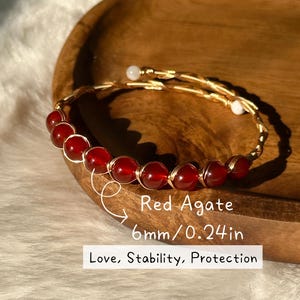 May include: A red agate bracelet with gold wire detailing. The bracelet features 6mm red beads and is described as promoting love, stability, and protection. The bracelet is displayed on a wooden tray.