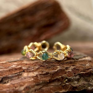 May include: A gold-colored ring with a series of colorful gemstones. The ring features a woven design with alternating pink, green, and clear stones. The ring is set against a textured, brown background.
