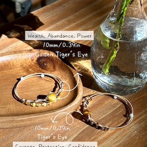 May include: Two golden bracelets with Tiger's Eye beads, each 10mm (0.39in) in diameter, are displayed on a wooden tray. The bracelets are next to a glass vase with a green stem. Text on the image reads: Wealth, Abundance, Power, Courage, Protection, Confidence.