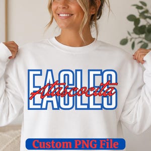 May include: A white long-sleeve sweatshirt featuring the word EAGLES in large blue outlined block letters. Overlaid across the center is the word Atascocita written in a red cursive script font. The bottom includes the text Custom PNG File.