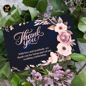 Op de afbeelding: Marineblauwe bedankkaart met ros&eacute;gouden belettering en bloemaccenten. De kaart luidt "Bedankt!" en bevat een boodschap van dankbaarheid voor een huwelijksfeest. De kaart is omgeven door groen en bloemen.