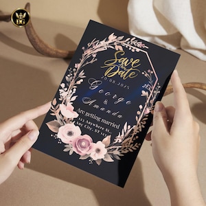 Op de afbeelding: Een zwarte trouwkaart met gouden tekst met de tekst "Save the Date" en de namen "George & Amanda". De uitnodiging is versierd met een bloemenkrans in roze en perziktinten.
