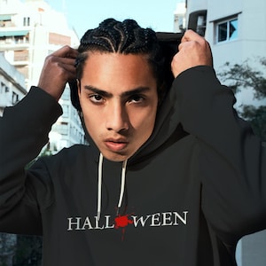 May include: A black hooded sweatshirt featuring the text HALL WEEN in white capital letters across the chest. Between the two words is a red, splattered graphic design. The hoodie includes white drawstrings hanging from the neckline.
