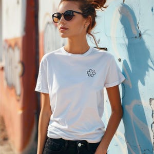 Op de afbeelding: Wit T-shirt met een kleine zwarte bloem opdruk. Het shirt heeft korte mouwen en een minimalistisch ontwerp. De persoon op de foto draagt een zwarte zonnebril en een zwarte jeans. De achtergrond is een kleurrijke graffiti muur.