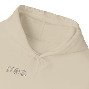 Op de afbeelding: Crèmekleurige hoodie met een trekkoordkap en een kleine afbeelding van drie schelpen in zwarte inkt. Het ontwerp omvat een kauri, een spiraalschelp en een sint-jakobsschelp, die een strandthema oproepen.
