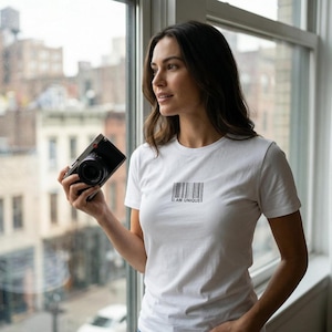 Peut inclure: T-shirt blanc avec le texte "I AM UNIQUE" imprimé sous un motif de code-barres. La personne tient un appareil photo noir. L'arrière-plan montre une vue de la ville à travers une fenêtre.
